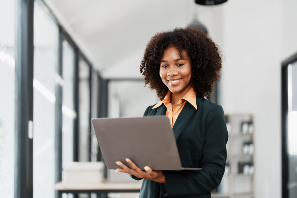 A young professional holding a laptop, standing in the middle of a modern office setting.